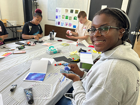 A student smiles at the camera while displaying her glass art as a table of students surrounded by painting tools focus on their work.