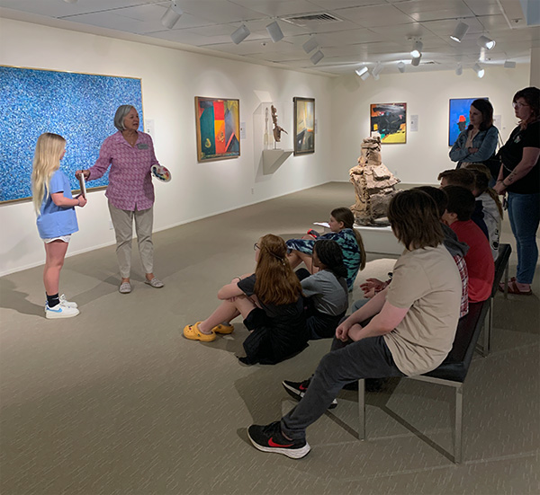 A docent stands in front of a seated group of schoolchildren, giving a tour of an art gallery.
